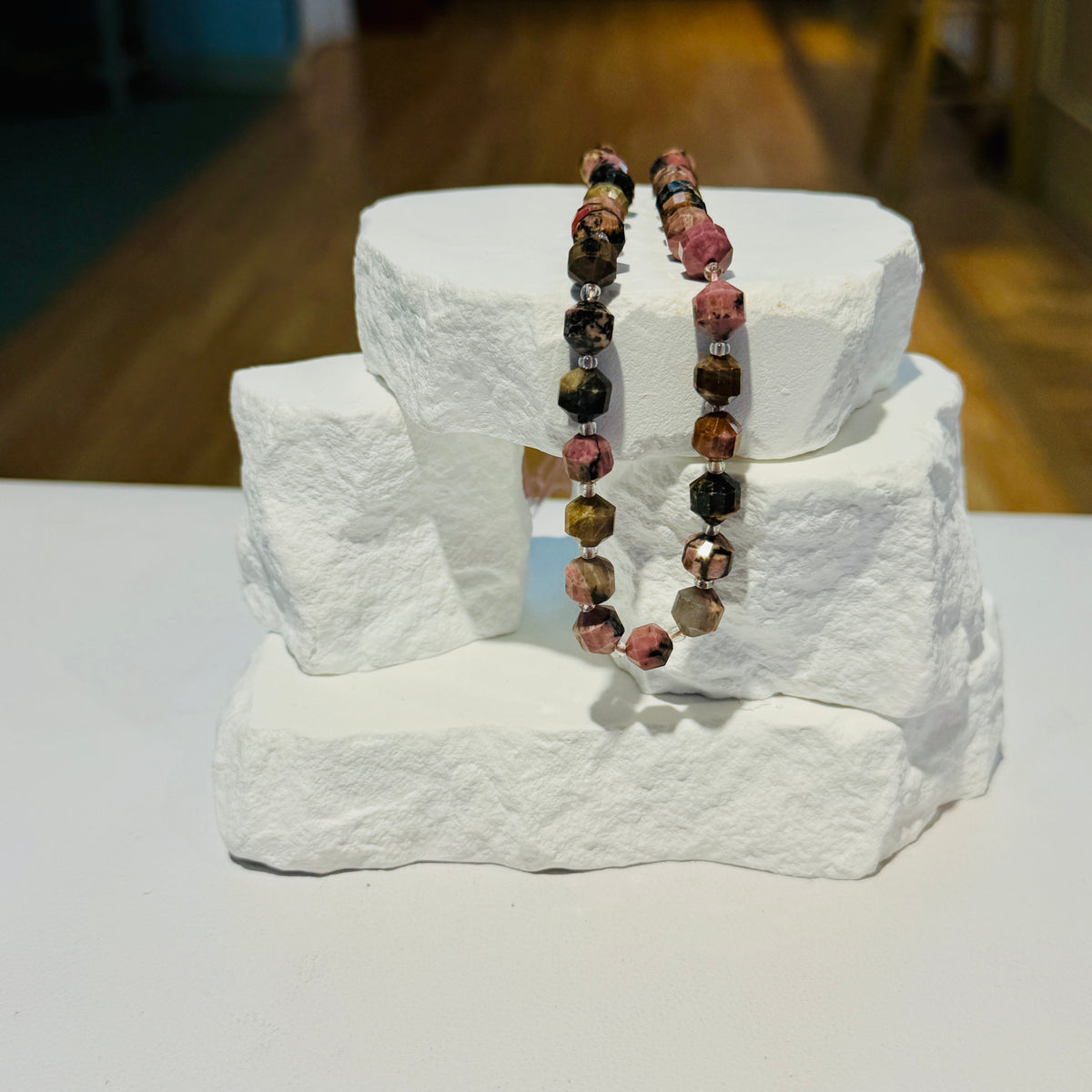 A strand of Rhodonite faceted round beads in pink, brown, black, and gold tones, draped over a white stone display stand on a white surface with a blurred wooden floor in the background.
