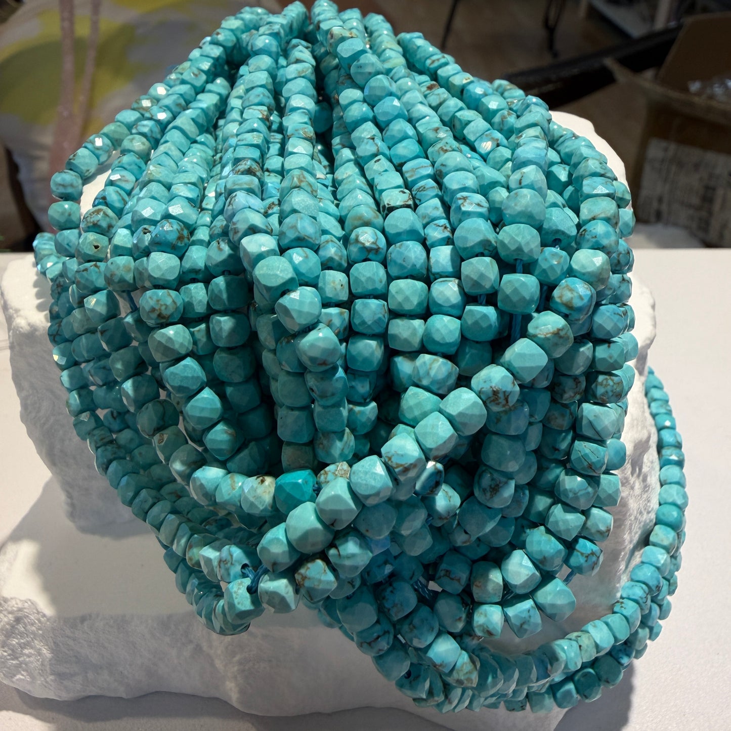 Multiple strands of faceted turquoise beads draped over a white display block, photographed indoors with a blurred background.