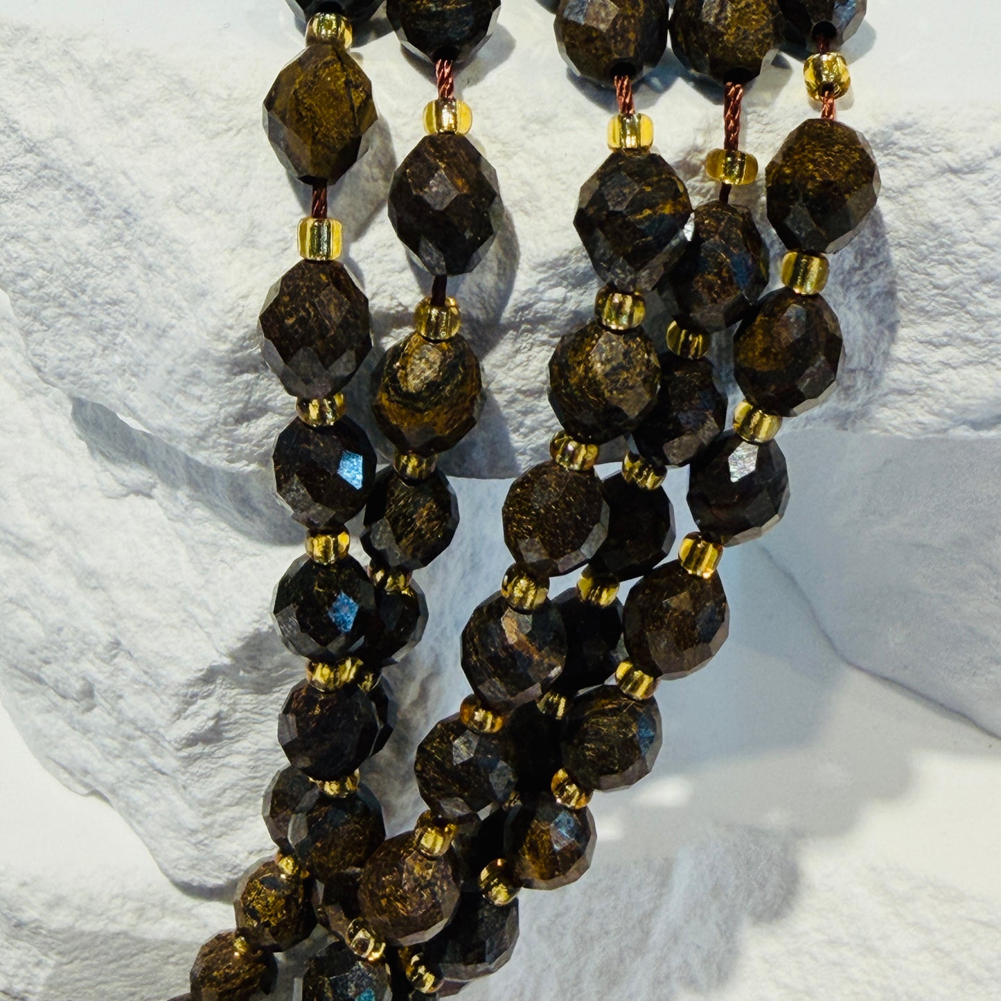 Multiple strands of faceted Bronzite beads separated by small gold spacer beads, draped over a white stone display block.