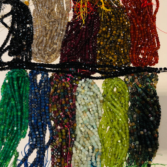 Multiple strands of faceted Gemstone Cube beads in various colors and patterns, arranged in vertical rows against a light background. 