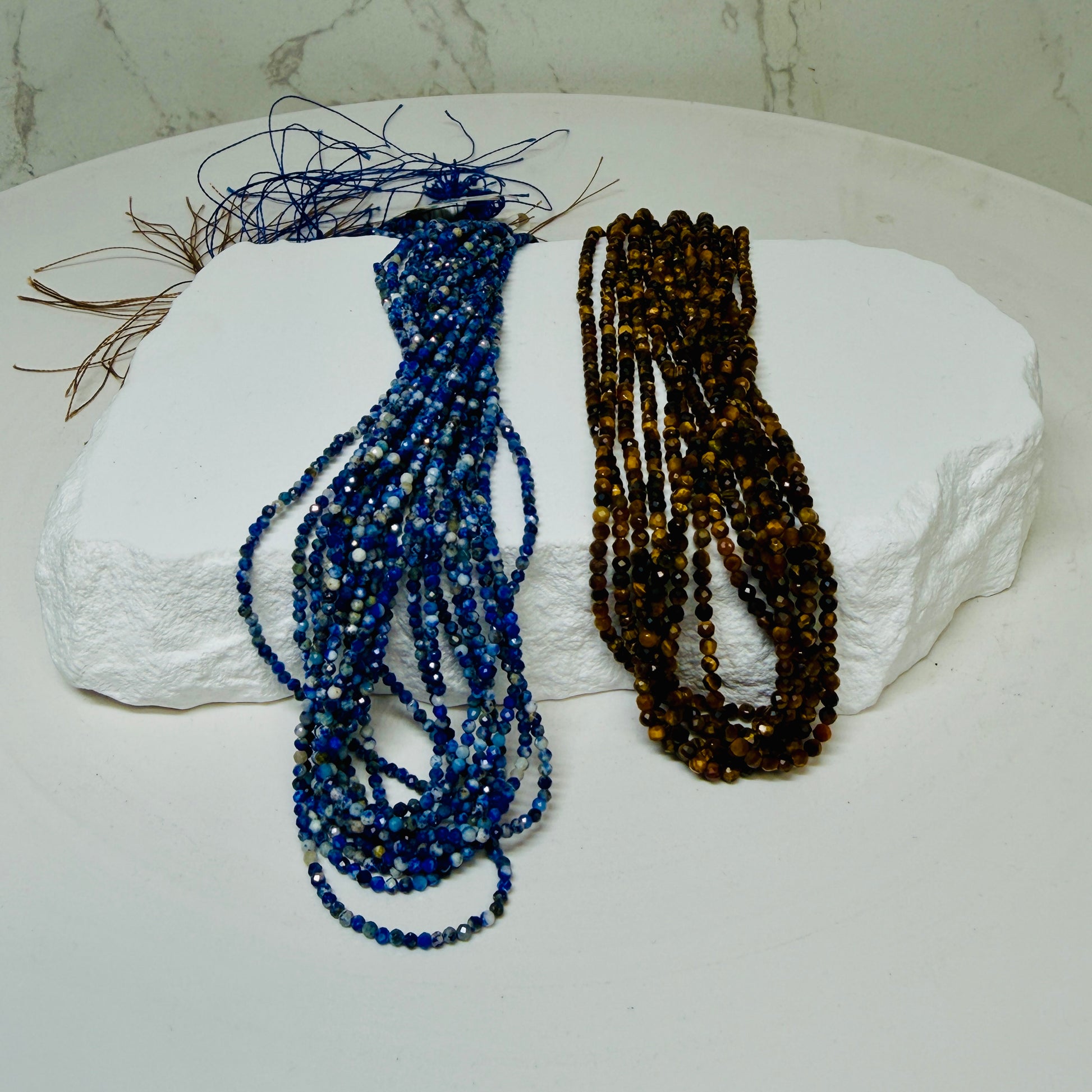 Small faceted lapis and Tiger eye bead strands draped over a white rock display on a white background