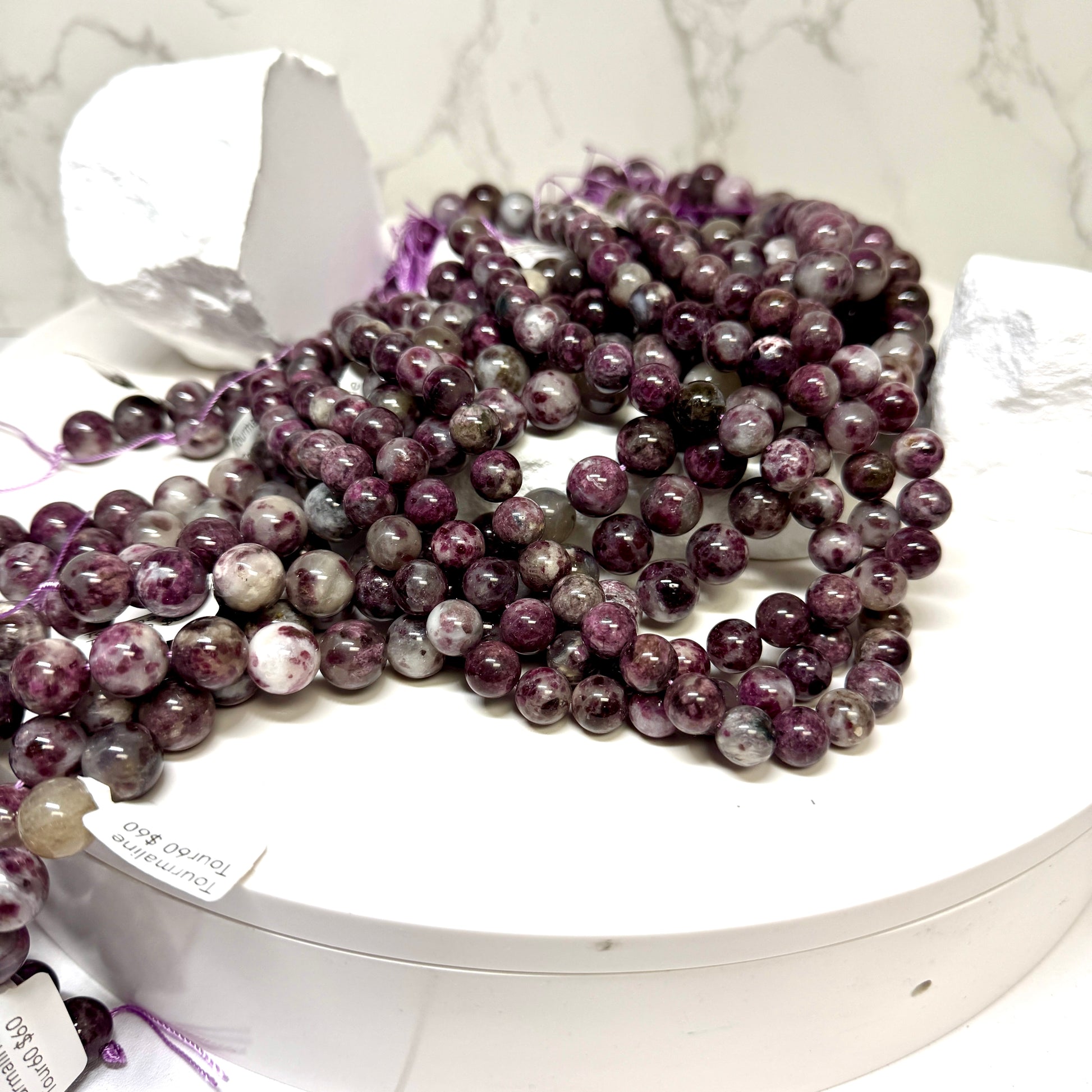 Multiple strands of polished round gemstone beads in shades of purple, pink, white, and gray, displayed on a white round tray with a white marble background.