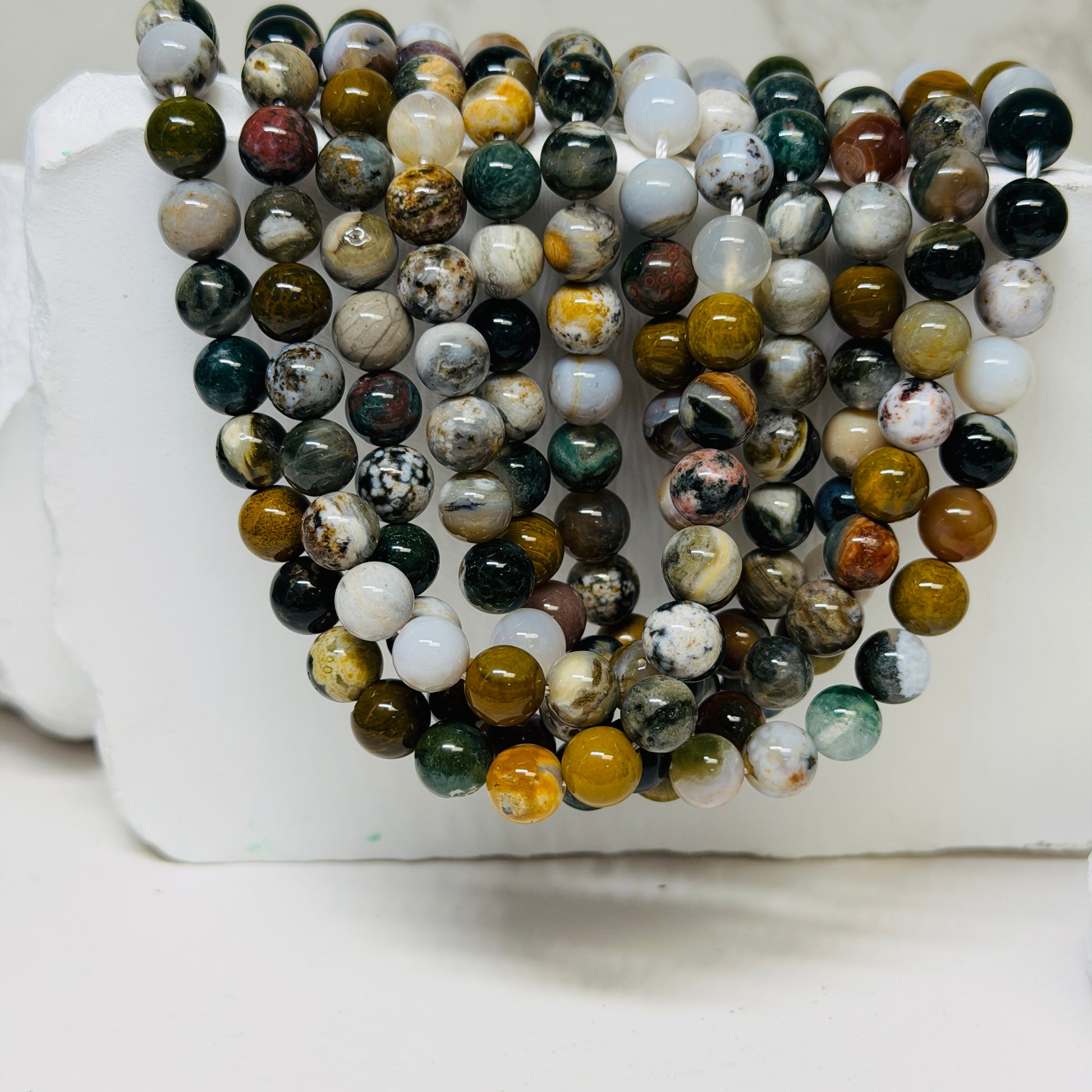 Multiple strands of polished round gemstone beads in assorted earthy colors and marbled patterns, draped over a white display block.