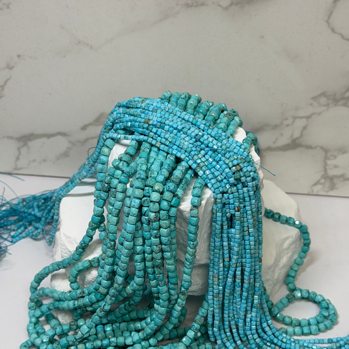 Multiple strands of faceted turquoise beads draped over a white display block, photographed indoors with a blurred background.