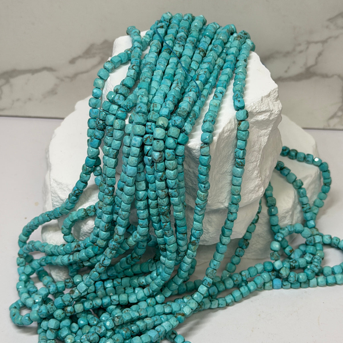 Multiple strands of faceted turquoise beads draped over a white display block, photographed indoors with a blurred background.