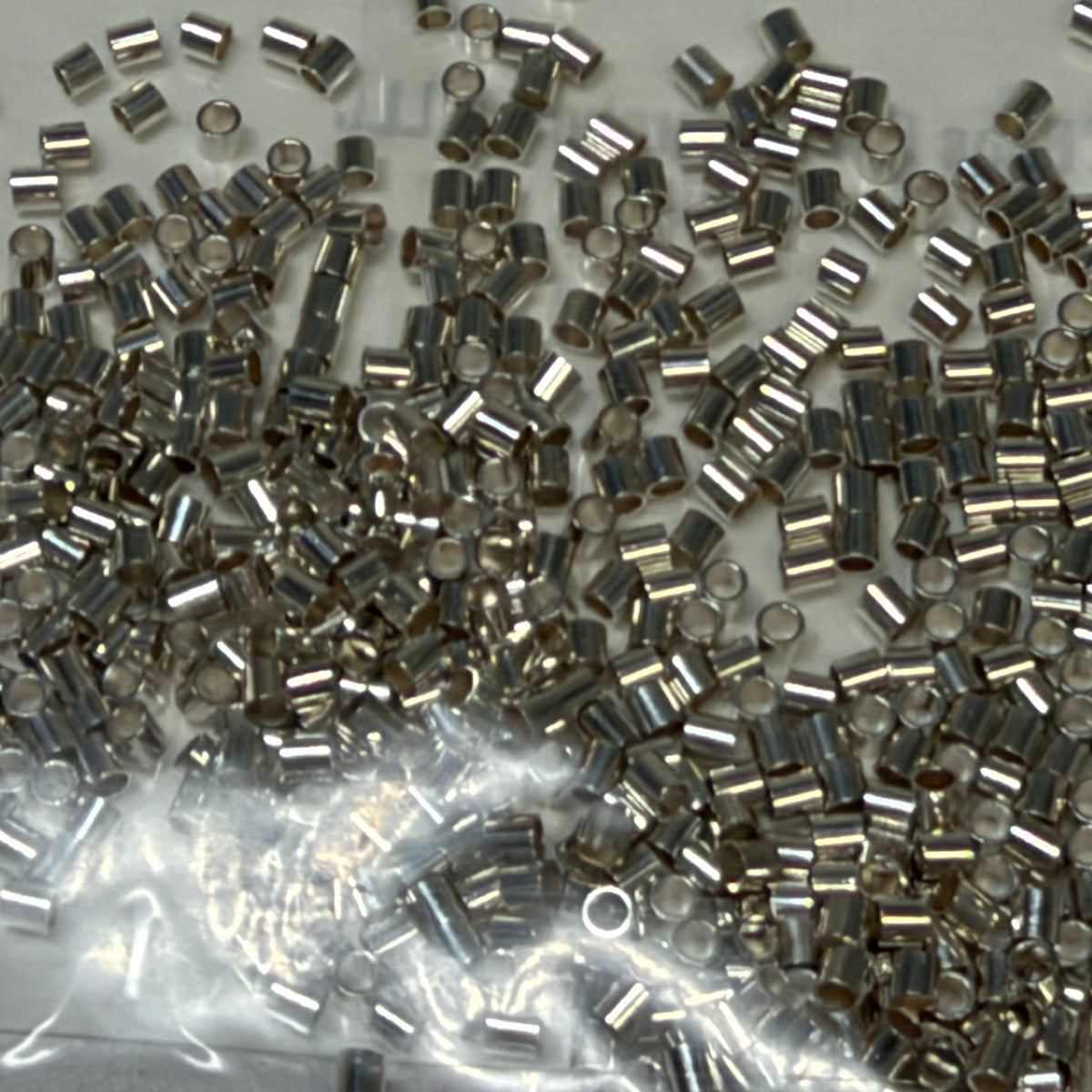 A pile of small Sterling Silver Crimp Beads with hollow centers, scattered on a light surface with a clear plastic bag visible in the lower portion.