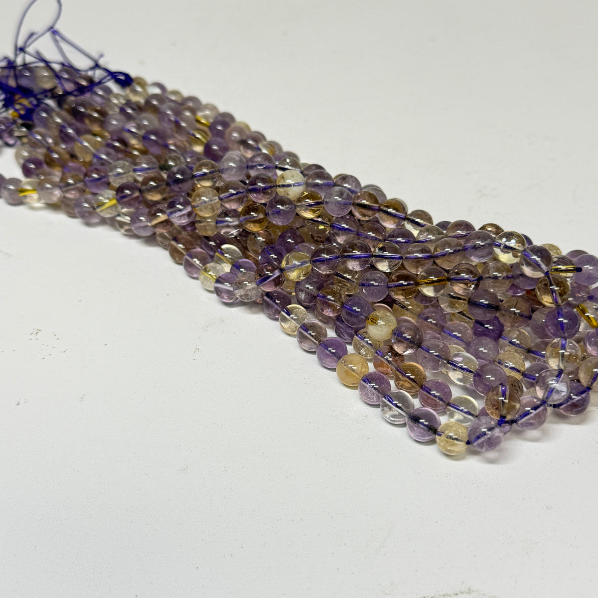 Multiple strands of faceted round gemstone beads strung on dark blue thread, arranged diagonally on a plain white surface. The beads show mixed colors including purple, brown, clear, and pale yellow.