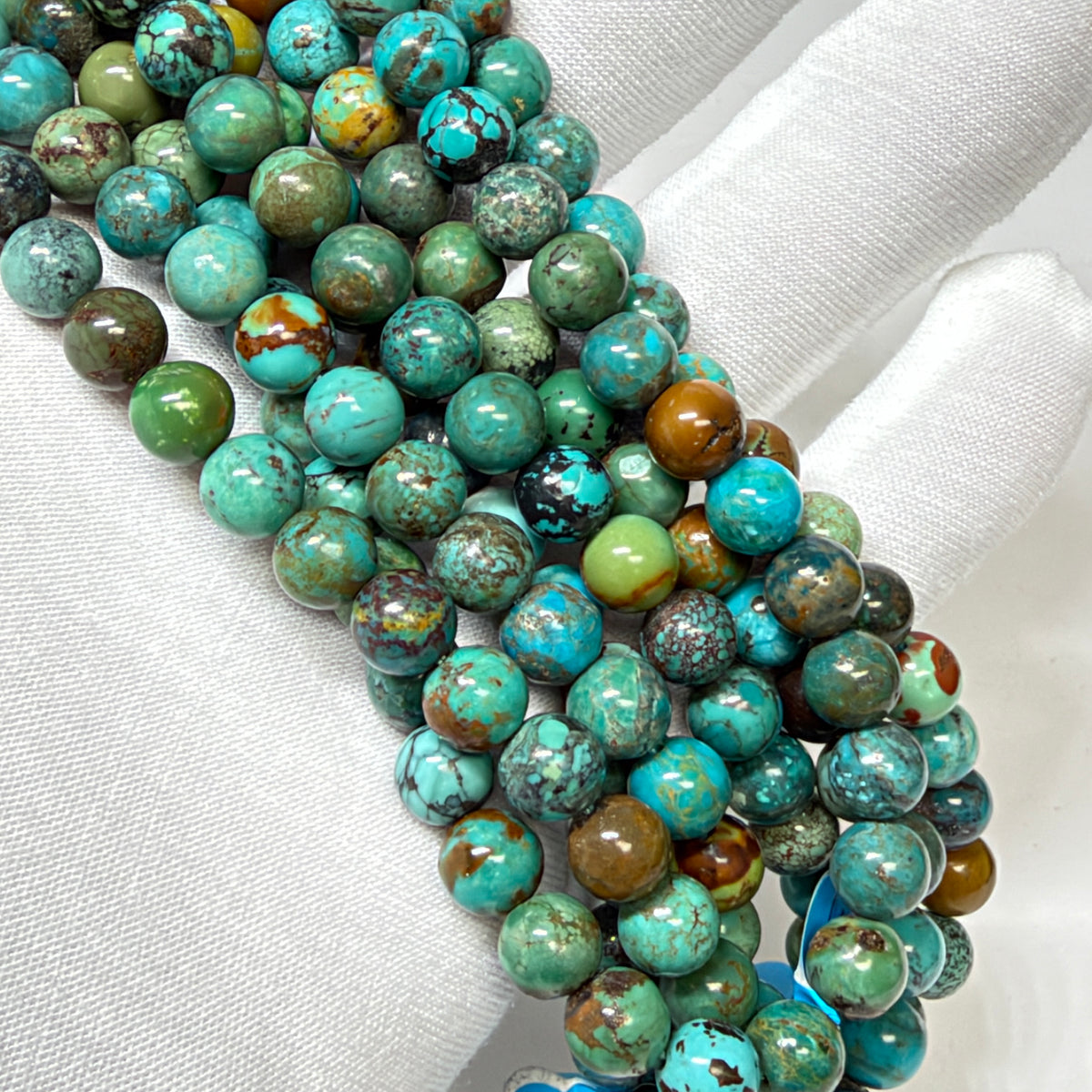 Multiple strands of polished round turquoise beads with brown and green matrix, displayed on a white fabric-covered hand.