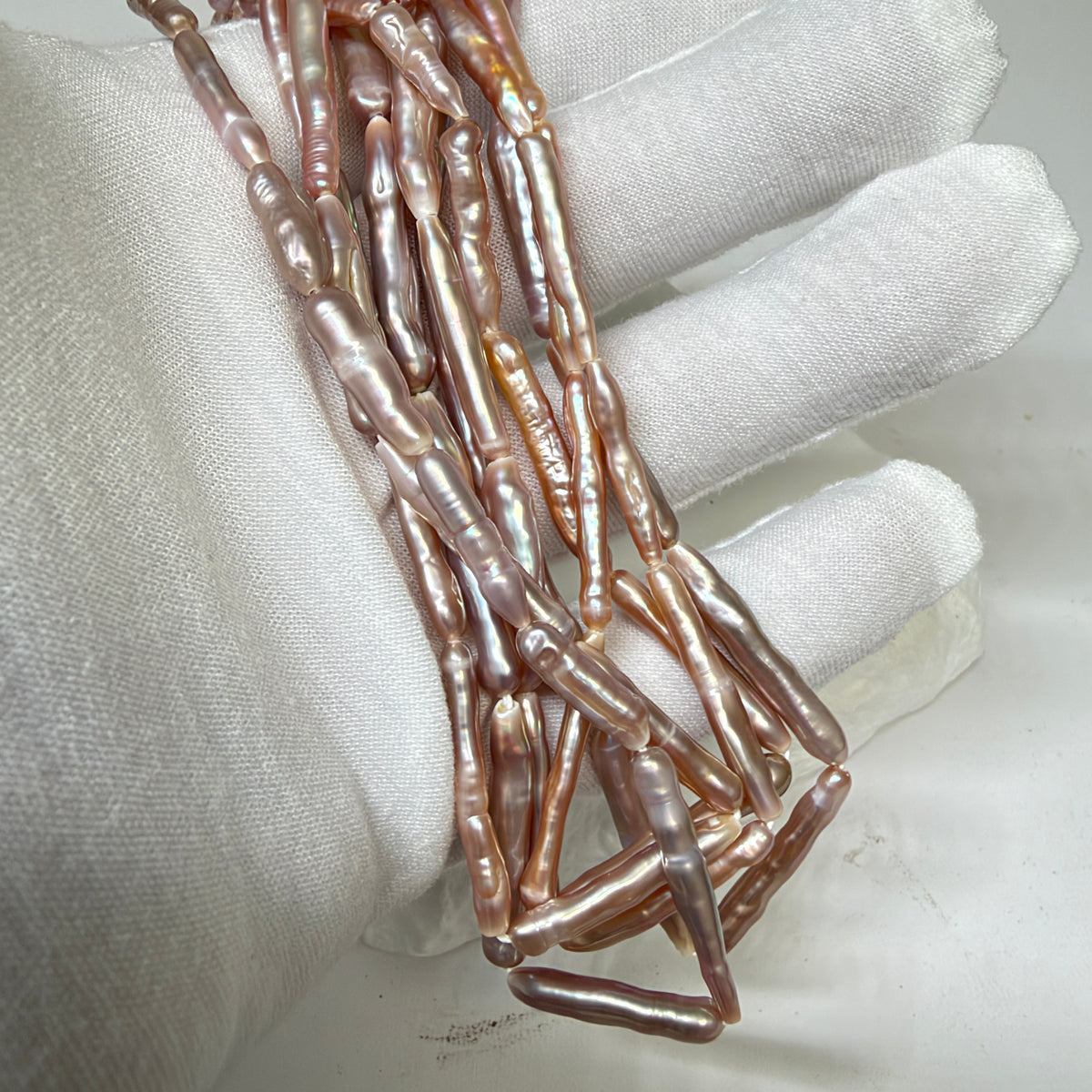 A close-up of a white fabric-covered hand holding a bundle of elongated, baroque-shaped pearl beads with a pearlescent rose-gold and purple luster.