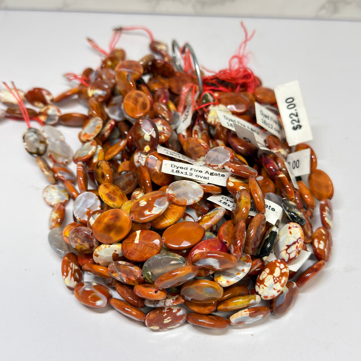 A tangled pile of strung, flat oval beads in mottled shades of reddish-brown, orange, and white. Small white tags attached to the red strings identify the beads as "Dyed Fire Agate 18x12 oval" and display a price of $22.00.