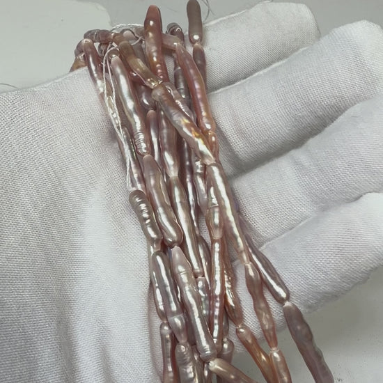 A close-up of a white fabric-covered hand holding a bundle of elongated, baroque-shaped pearl beads with a pearlescent rose-gold and purple luster..  This is a video