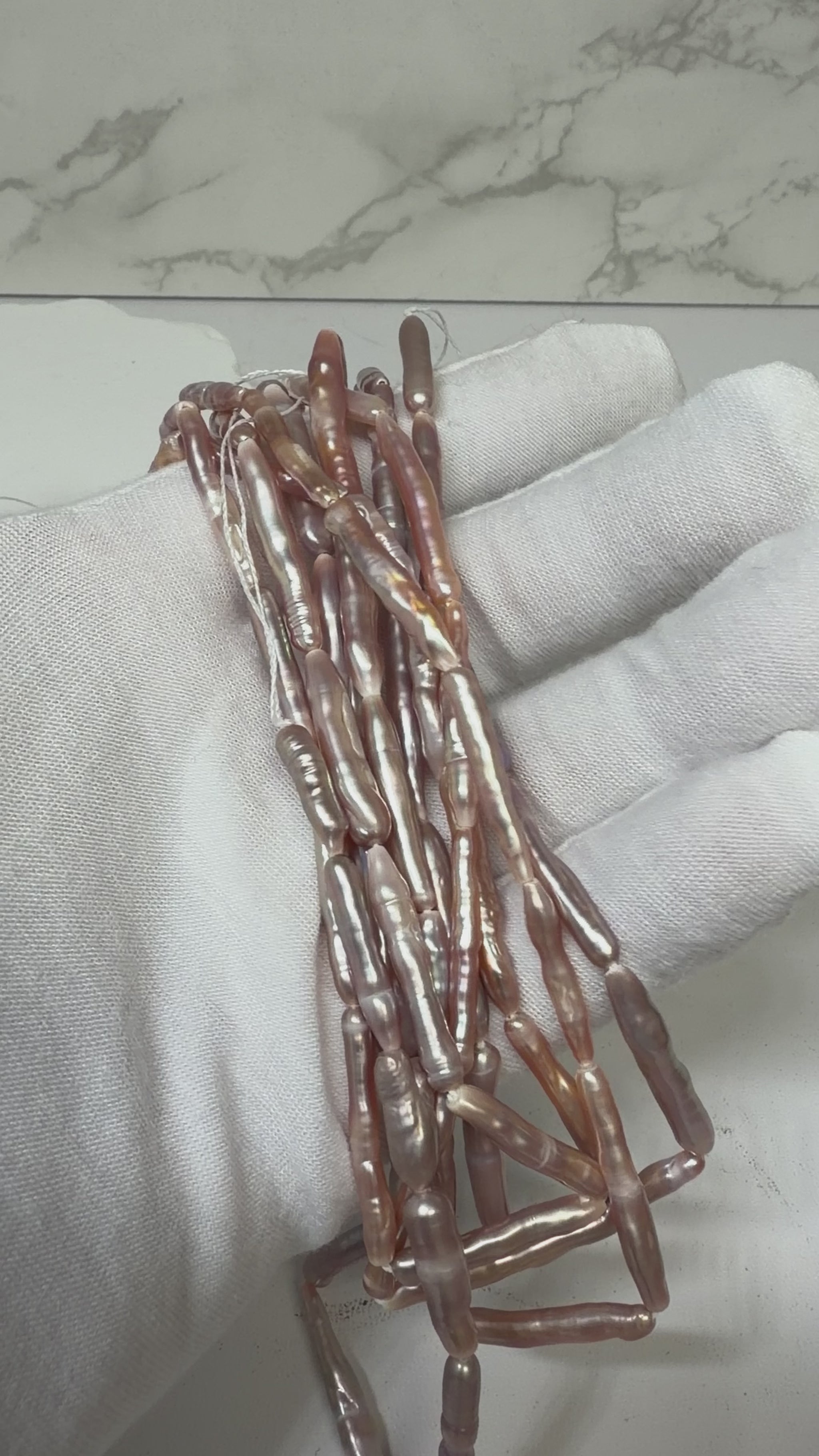 A close-up of a white fabric-covered hand holding a bundle of elongated, baroque-shaped pearl beads with a pearlescent rose-gold and purple luster..  This is a video